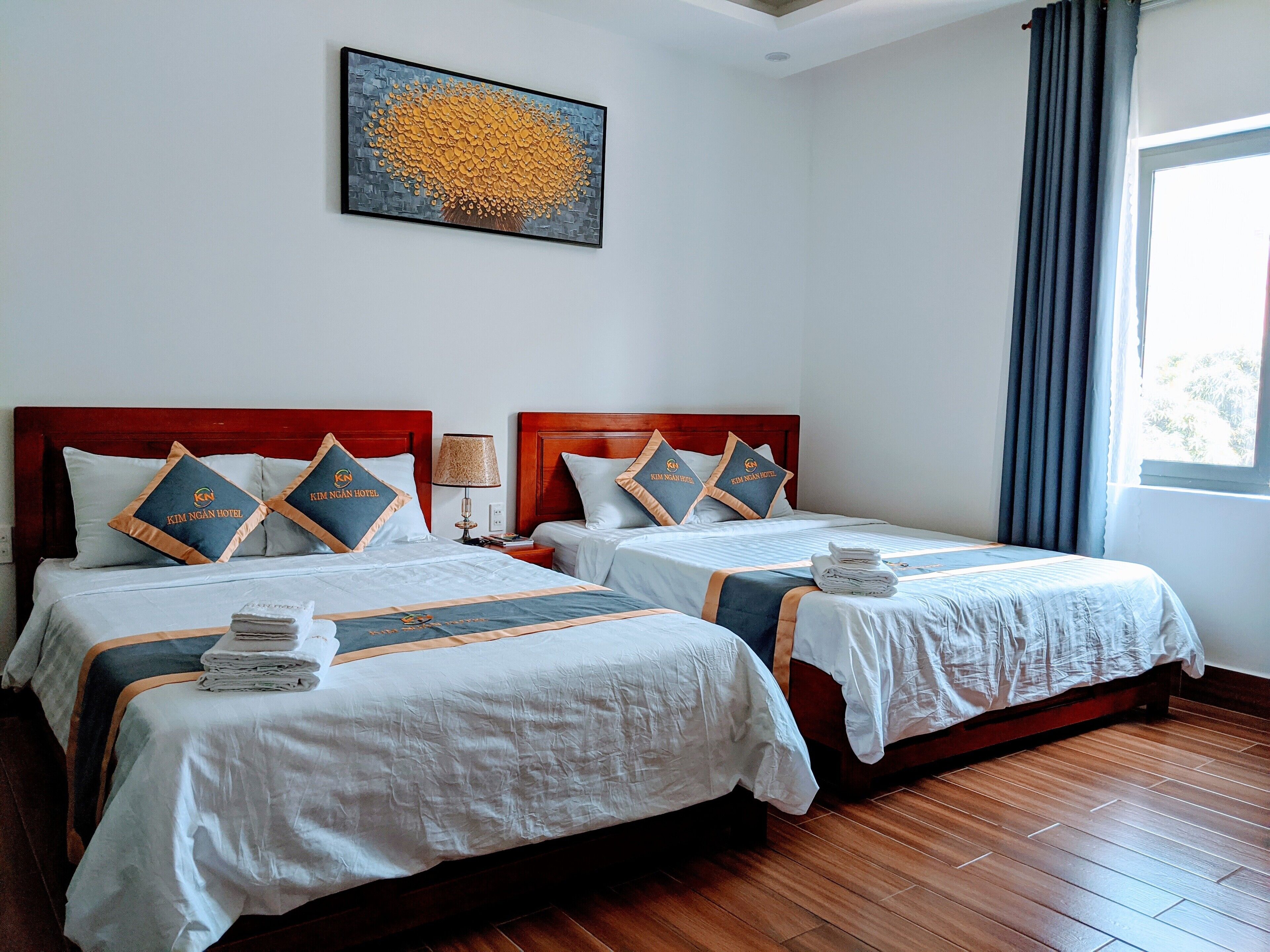A hotel room with two beds, each with matching headboards and pillows, a nightstand with a lamp, and a window with curtains.