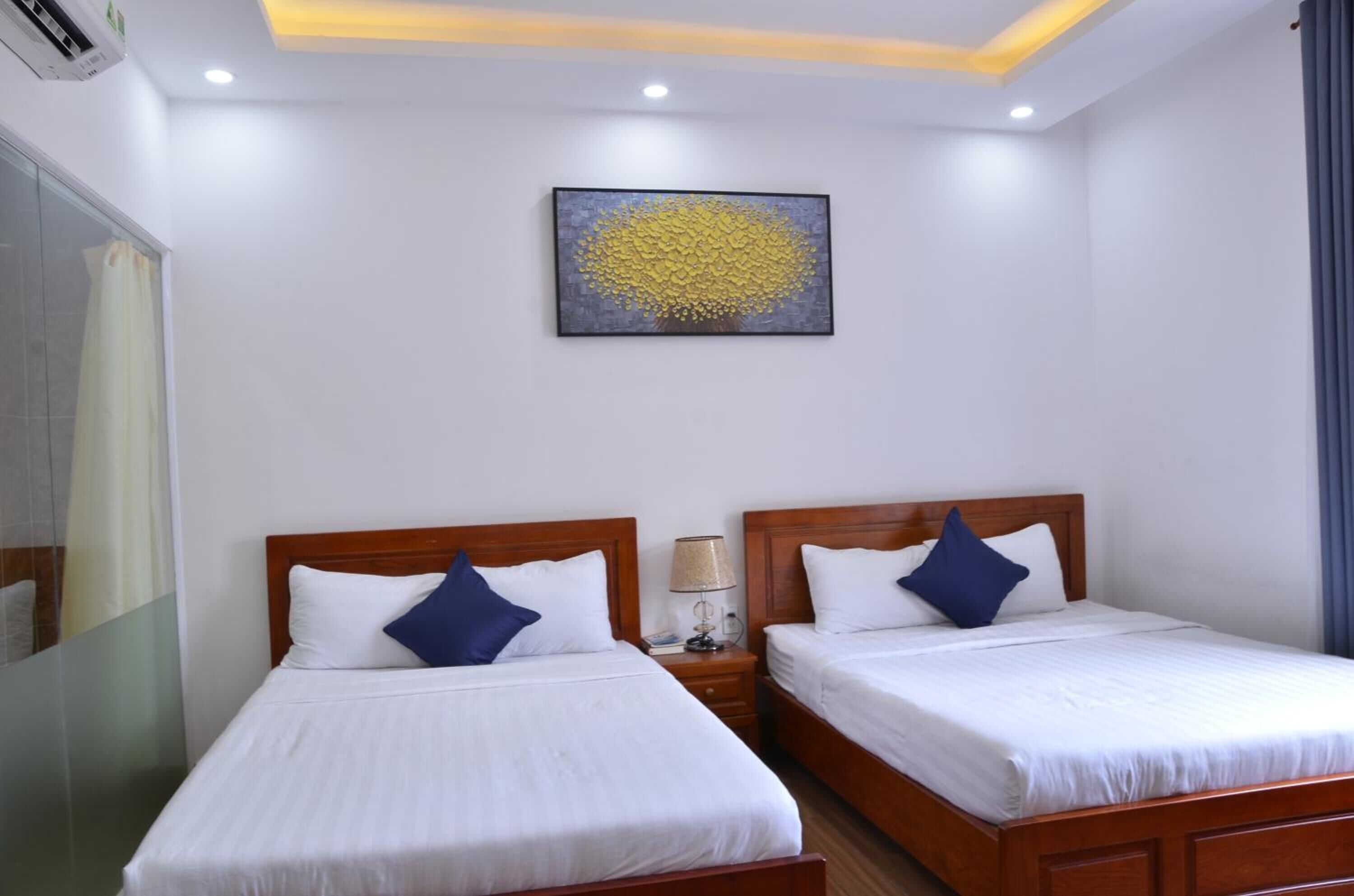 Habitación de hotel con dos camas, una lámpara de techo, una obra de arte en la pared y una mesita de noche con lámpara.