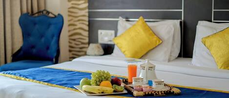 A hotel room with a neatly made bed, a tray of fruits and juice, and a breakfast setup.