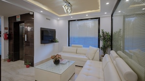 A modern living room with a glass coffee table, white sofas, a flat-screen TV, and a large window with blinds. - BlueNeck Sec 40 Unitech Cyber Park Gurugram (Gurugram)