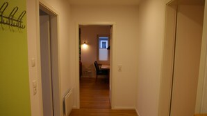 A narrow hallway with wooden flooring, a dining area in the background, and a door on the left.