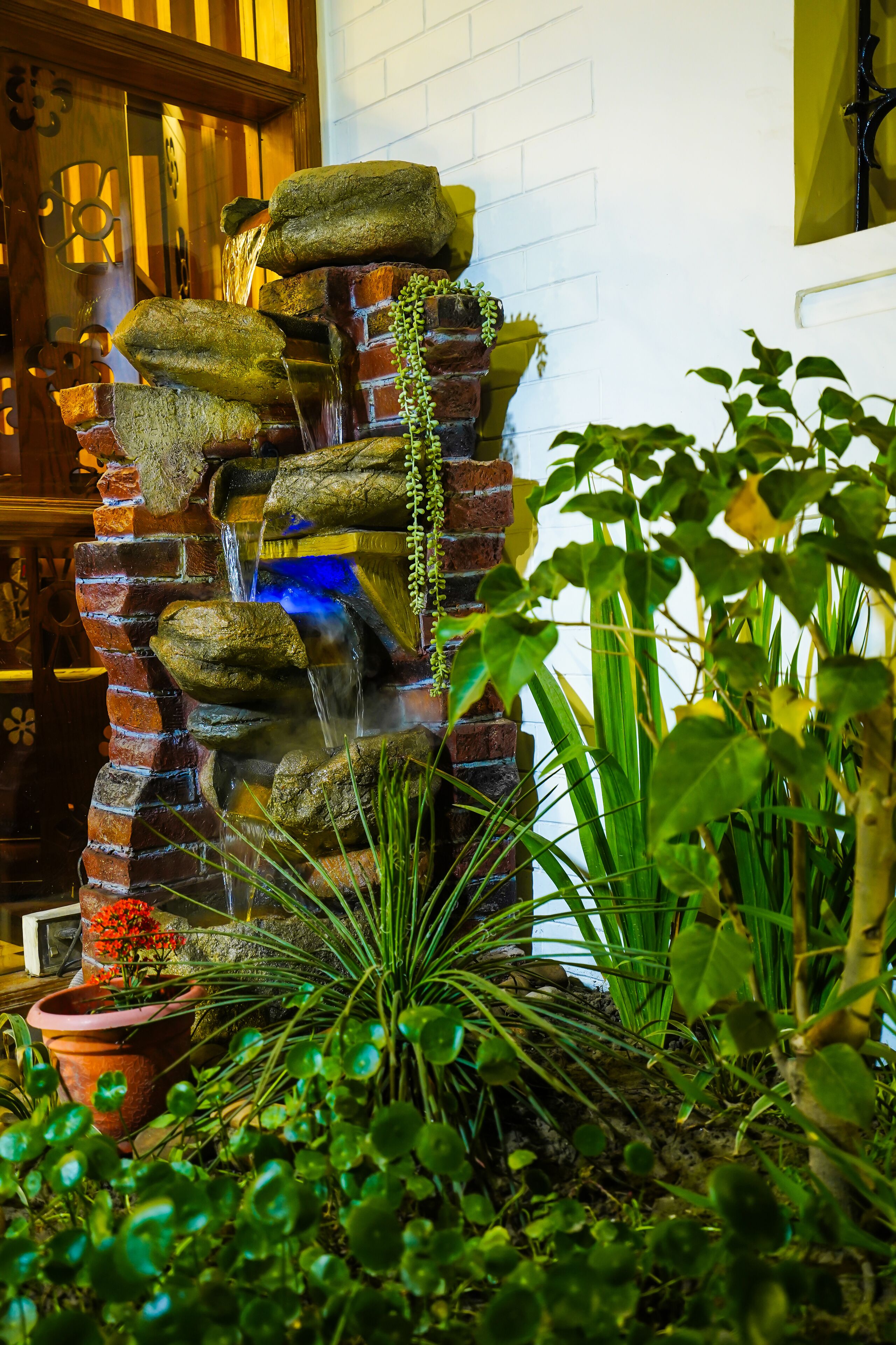A brick waterfall feature with cascading water, surrounded by various plants and a potted plant with red flowers.