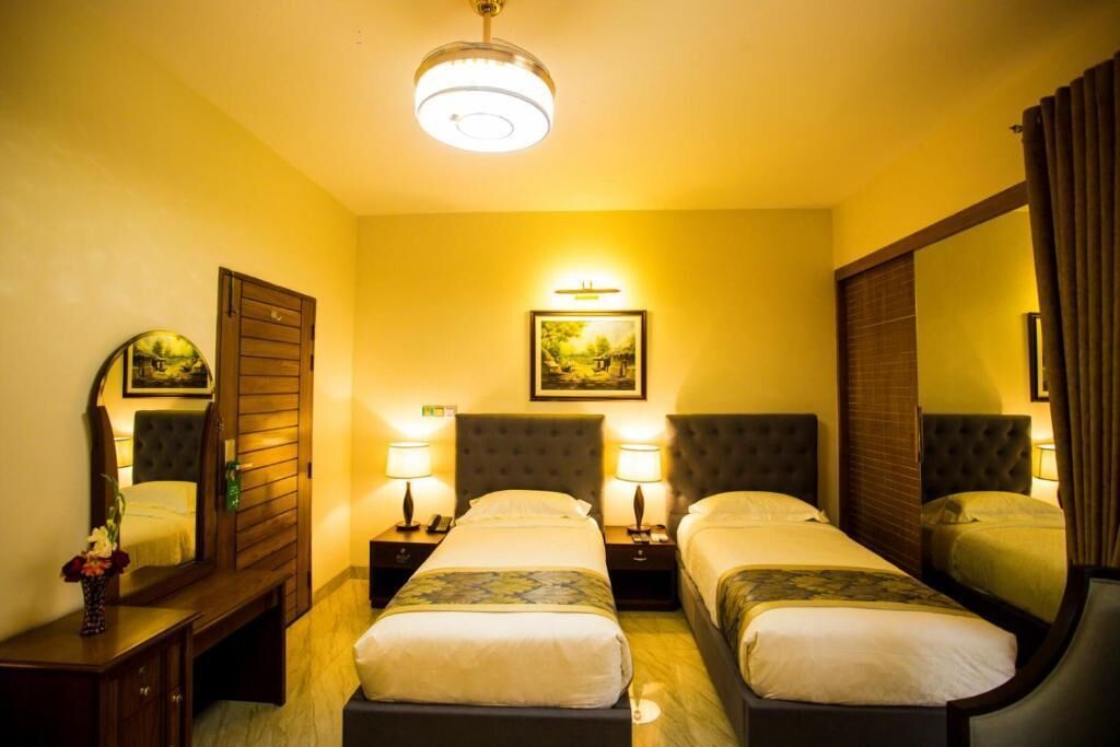 A hotel room with two single beds, a large mirror, a nightstand with a lamp, and a framed picture on the wall.