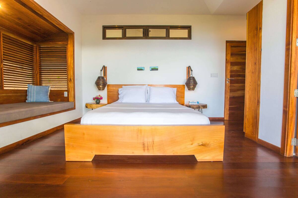 A large wooden bed with a headboard, wooden flooring, and a bench with a towel.