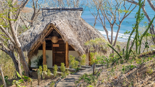 A thatched-roof hut with a wooden door and a small porch, surrounded by cacti and trees, overlooking a beach. - Verdad Nicaragua Beach Hotel & Retreat (San Juan del Sur)