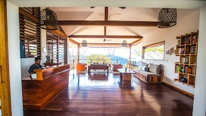 A spacious lobby with a wooden reception desk, a seating area with a sofa and chairs, a bookshelf, and a view of the outdoors. - Verdad Nicaragua Beach Hotel & Retreat (San Juan del Sur)