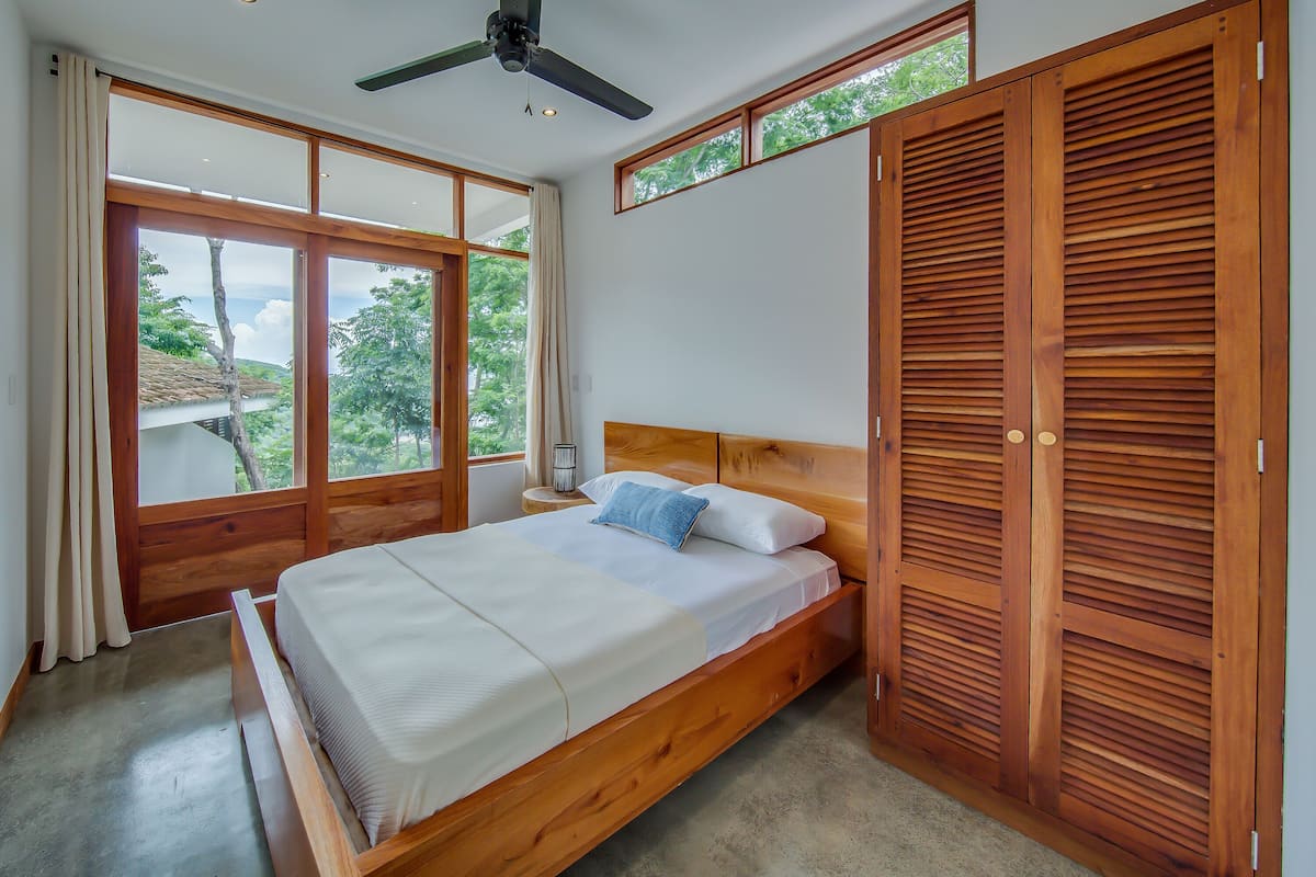A bedroom with a wooden bed, a ceiling fan, and large windows offering a view of the outdoors.