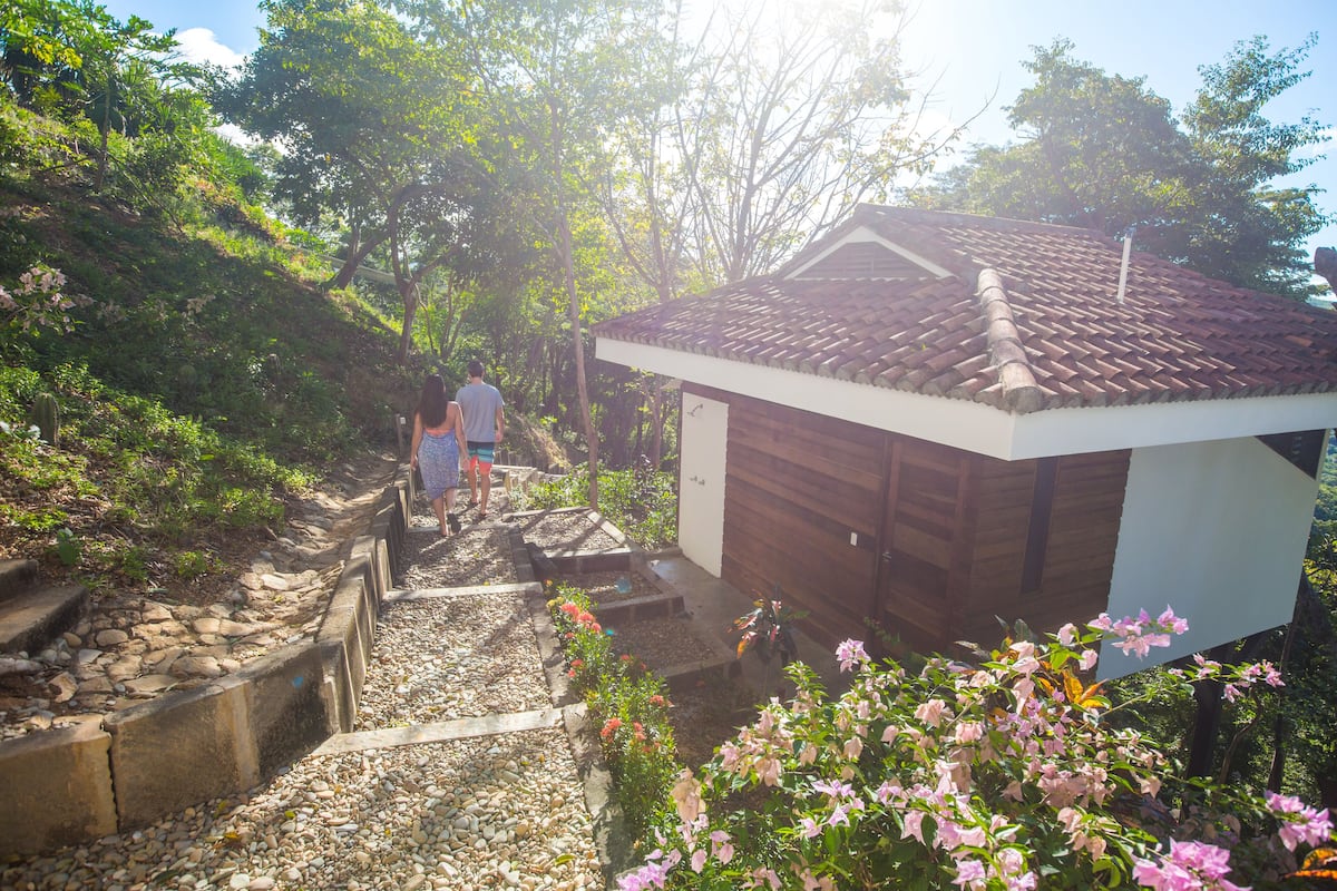A small wooden cabin with a tiled roof, surrounded by a garden with various plants and flowers.