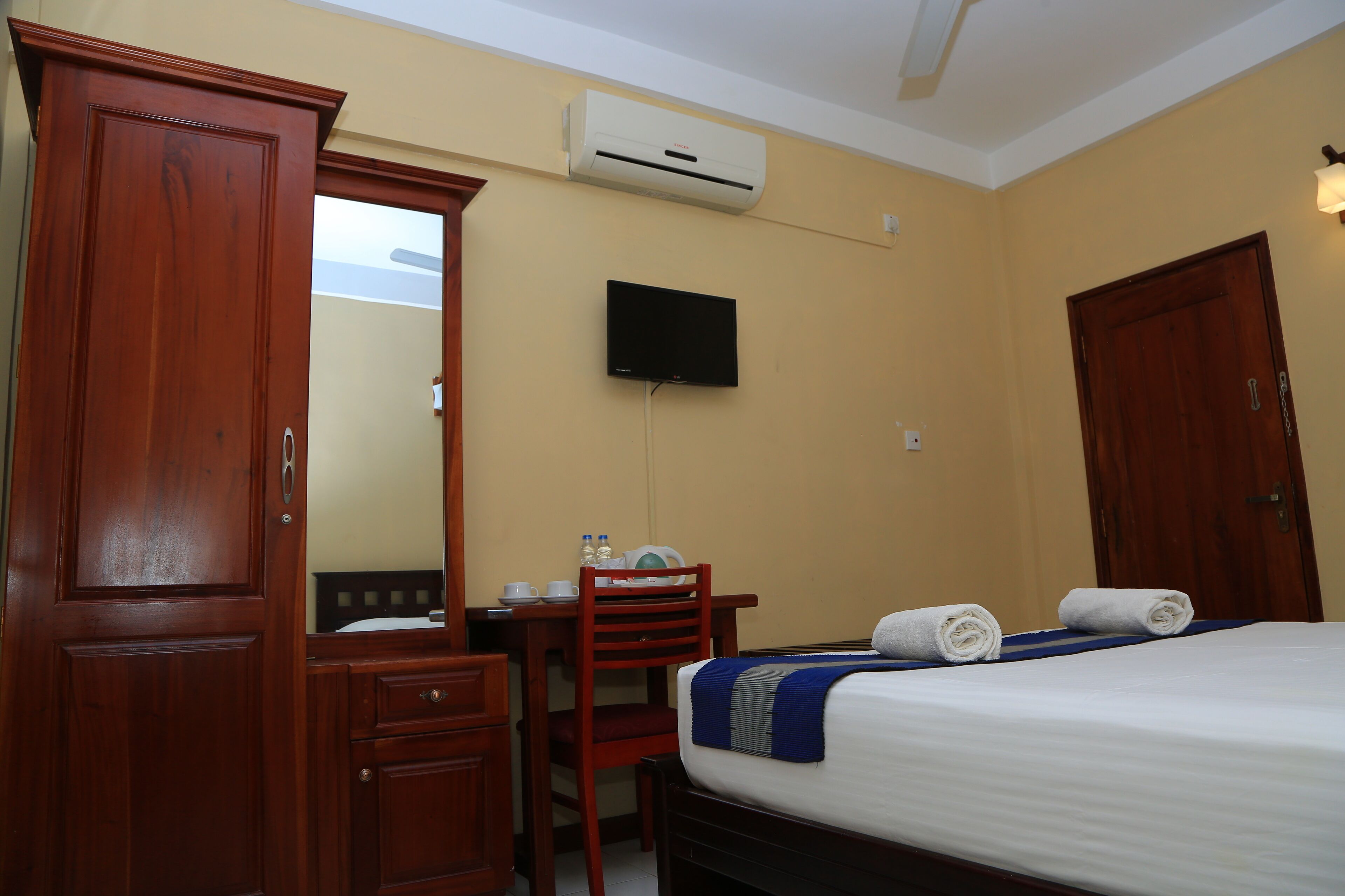 A hotel room with a bed, a desk with a chair, a wardrobe, a television, and an air conditioning unit.