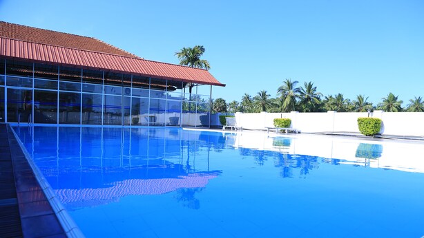 A clear blue pool with a tiled deck, a red-roofed building, and palm trees in the background. - PJ Hotels Jaffna (Valikamam East)