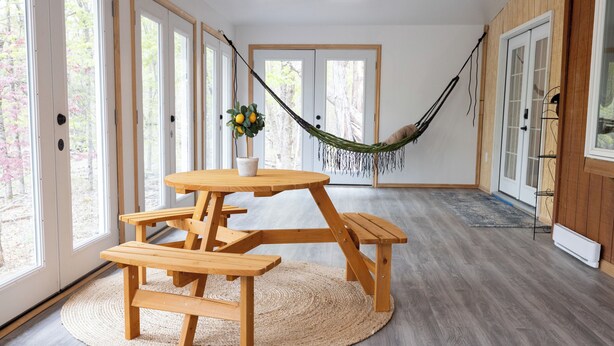 Una mesa de picnic de madera con bancos, una hamaca y una planta en maceta en una habitación con ventanas amplias y suelo de madera.