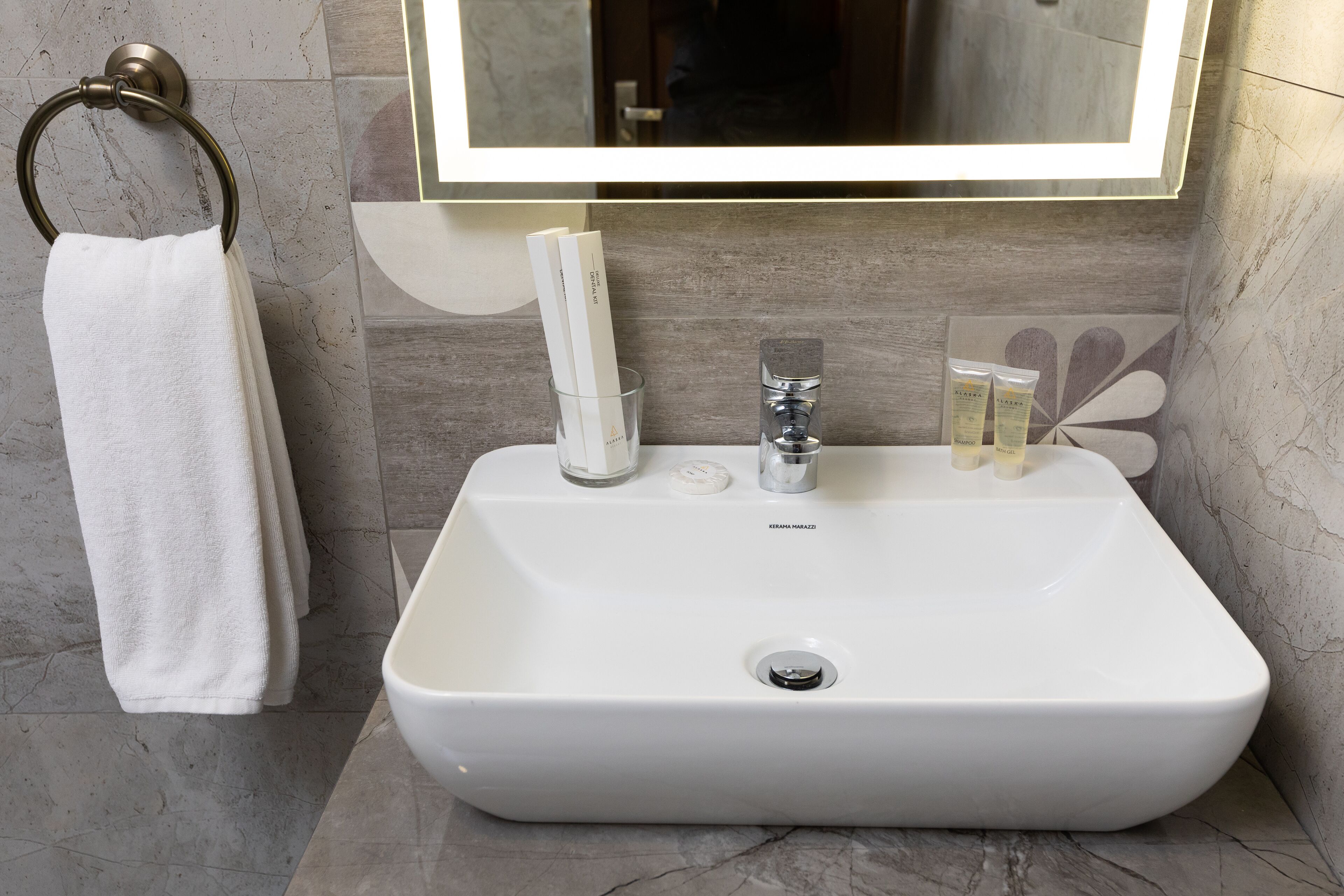 Un baño con lavamanos blanco, argolla para toallas y espejo con luz superior.