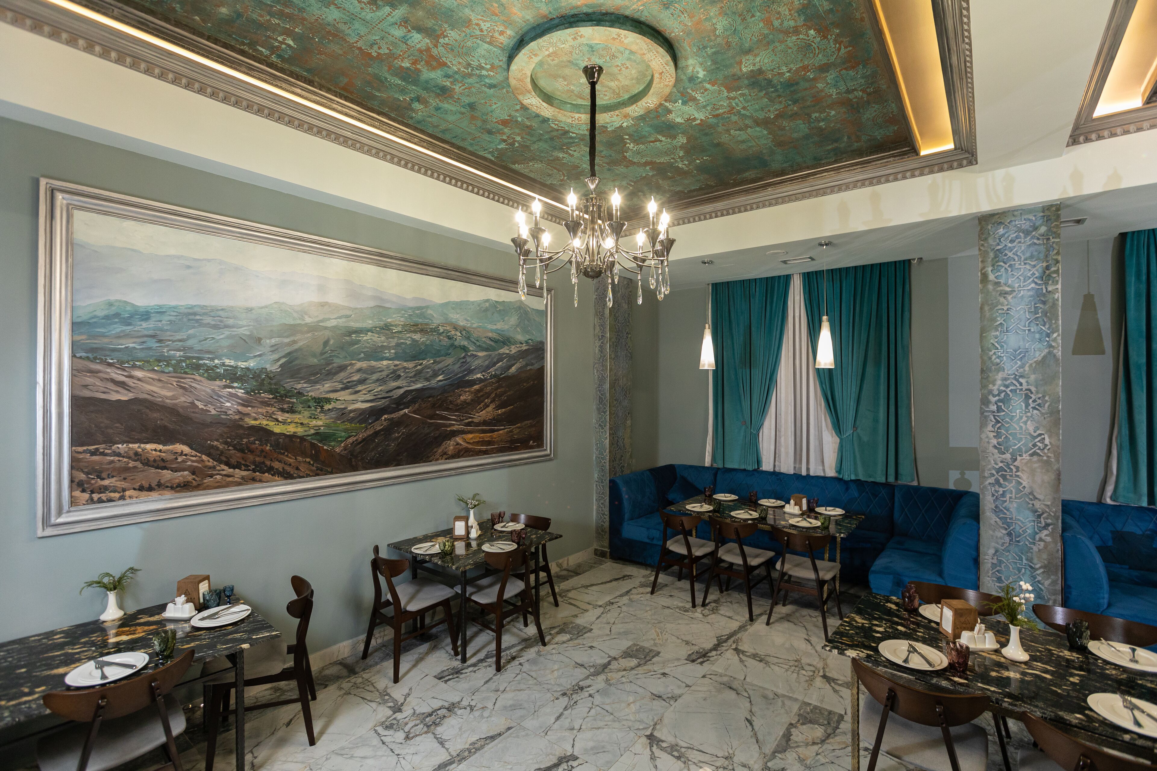 A dining area with a large landscape painting, a chandelier, and blue upholstered seating.