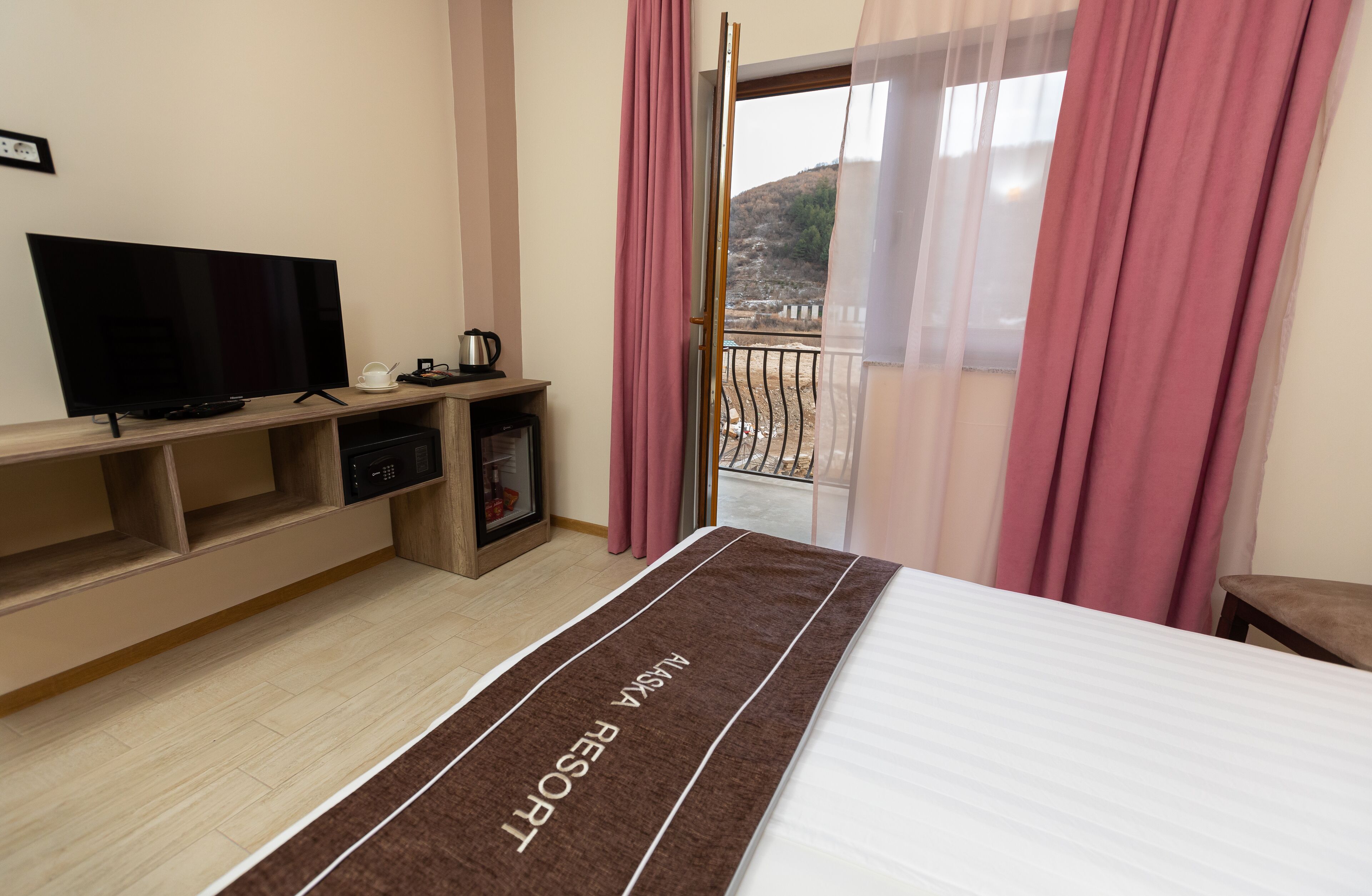 A hotel room with a bed, a TV, a small table with a kettle, and a balcony with pink curtains.