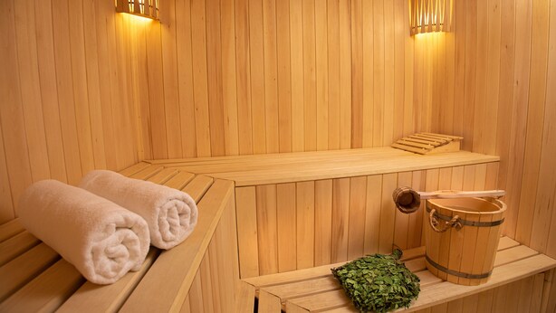 Una sauna de madera con bancos, un banco con toallas y un cubo de hojas.