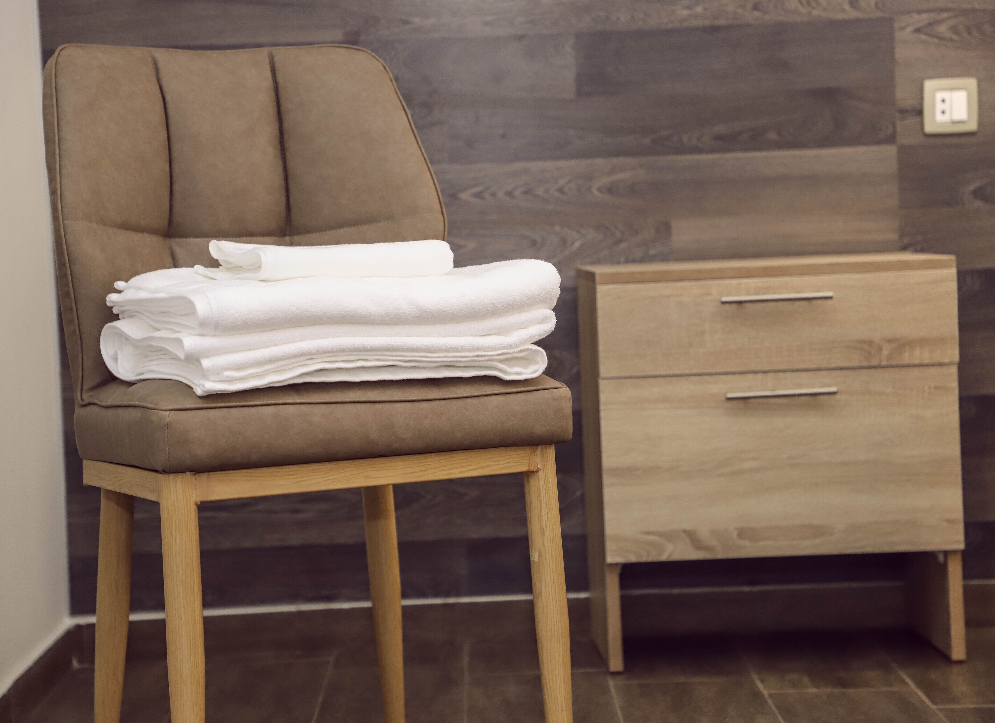 A brown upholstered chair with a stack of white towels, a wooden nightstand with two drawers, and a dark wood-patterned wall.