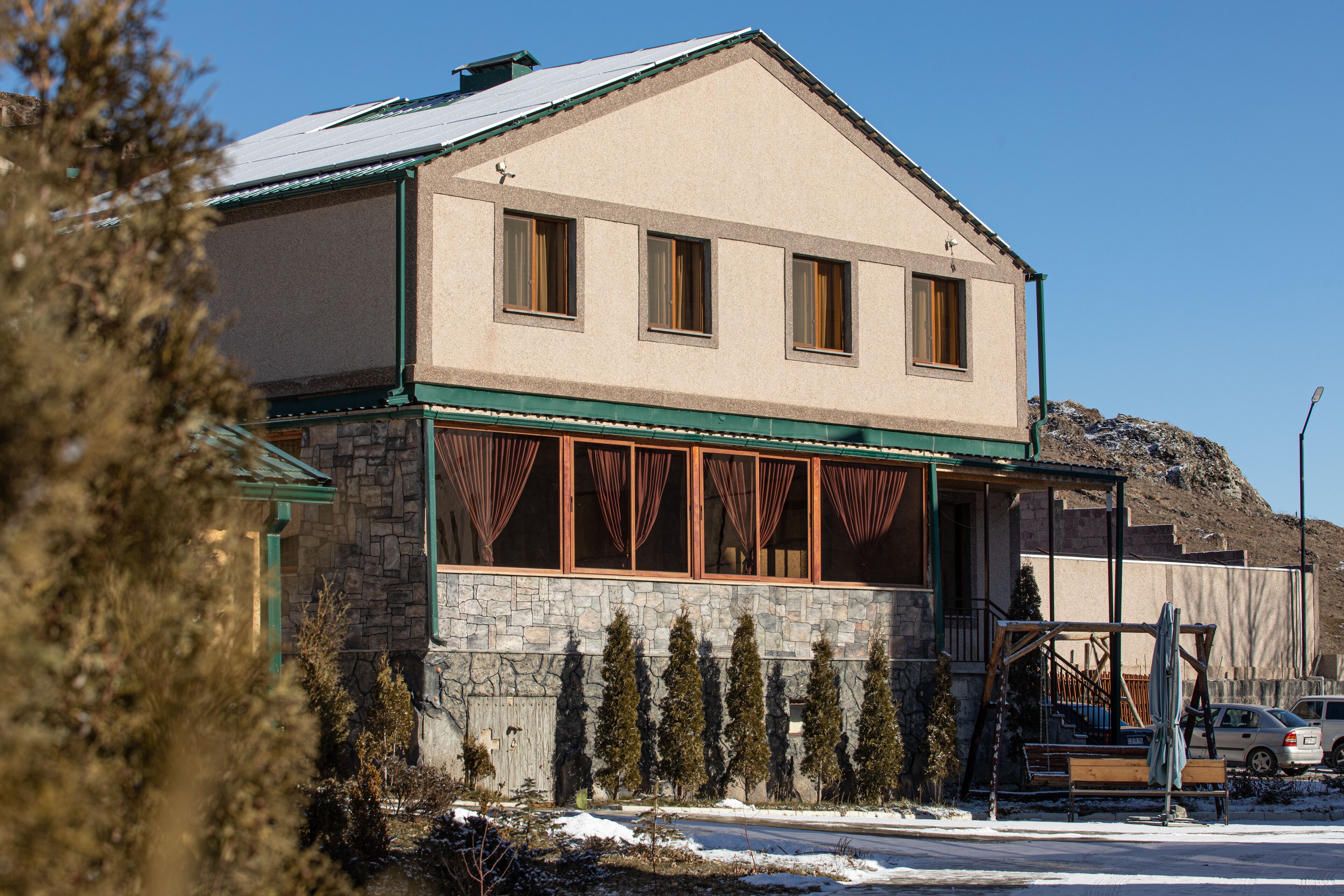 Un edificio de dos pisos con fachada de piedra y detalles verdes, que incluye un porche con barandas de madera y un banco.