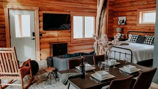 A log cabin interior with a dining area, a sofa, a television, a fireplace, and a bed.