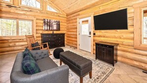 A log cabin interior with a sofa, ottoman, wooden dresser, and a flat-screen TV mounted on the wall.