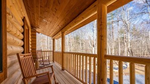 A log cabin with a wooden porch, wooden chairs, and a view of a forest.