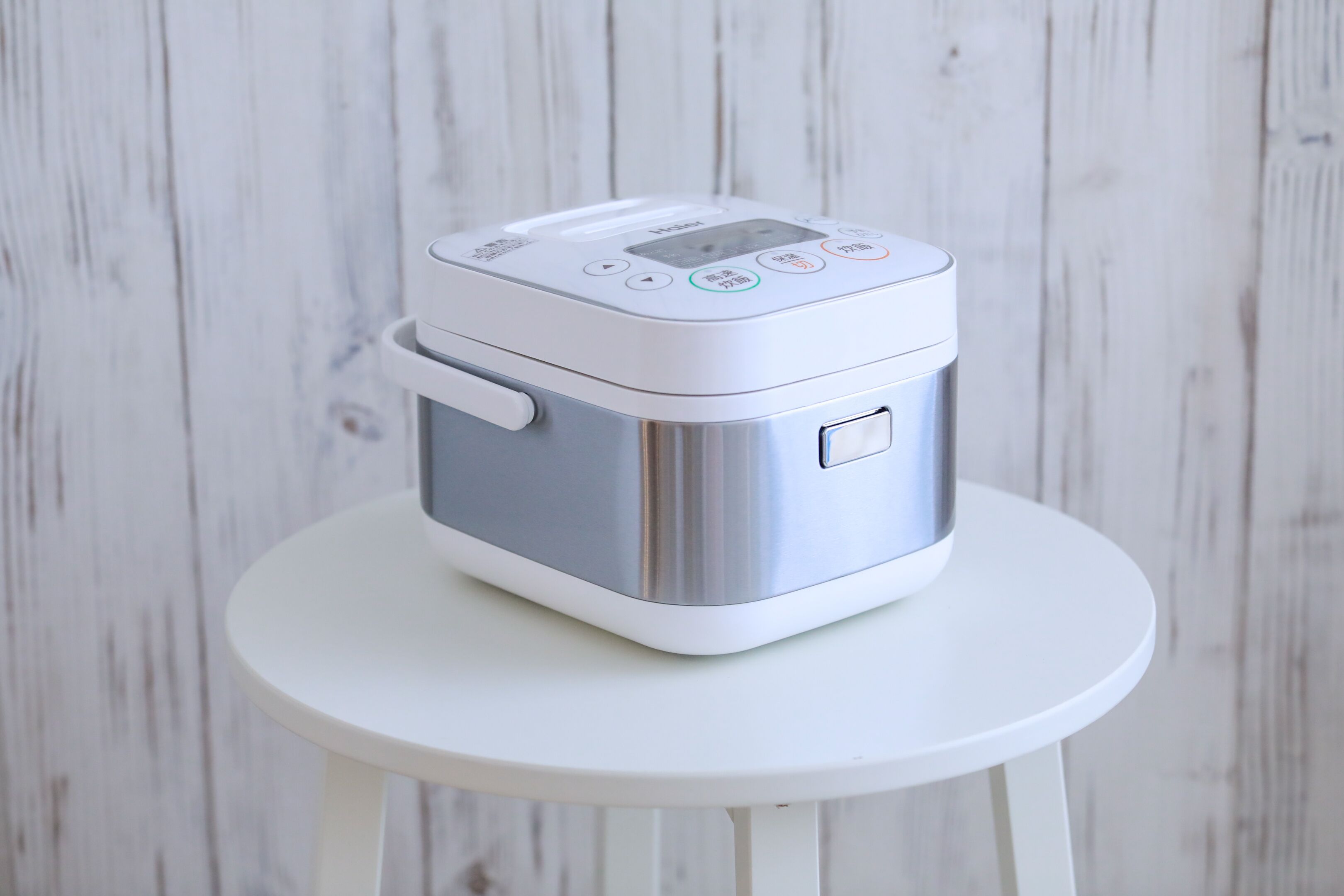 A stainless steel and white electric rice cooker on a white stool against a white wooden wall.