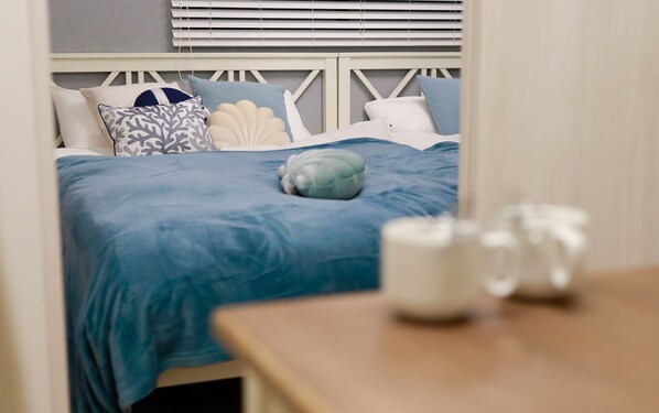 A neatly made bed with a blue bedspread and decorative pillows, a wooden nightstand with a white mug, and a window with blinds in the background.