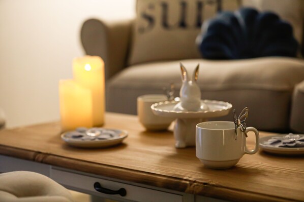 A cozy living room with a wooden coffee table, a lit candle, a white teapot, a teacup with a butterfly decoration, and a sofa with a cushion.