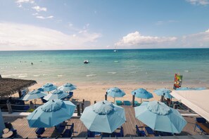 Zona de playa con sombrillas azules y sillas de descanso, una terraza de madera y vistas al océano.