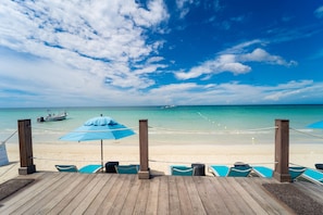 A wooden deck with blue umbrellas and chairs overlooking a beach with a boat in the distance. - Blue Skies Beach Resort (Negril)