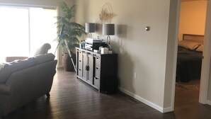 A spacious living room with hardwood flooring, a sofa, a cabinet with a flat-screen TV, and a large window providing natural light. - 12 minutes to PennState (Bellefonte)