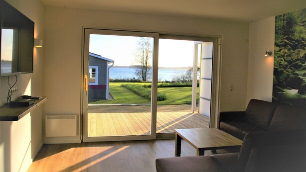 A modern room with a sliding glass door leading to a deck with a view of a house and a lake. - Vacation home H (77 m²) with lake view for up to 4 people (Klein Wittensee)