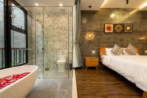 A modern bathroom with a glass shower enclosure, a bathtub with red rose petals, and a wooden bedside table with a bed. - Paddy Boutique House Hoian (Da Nang)