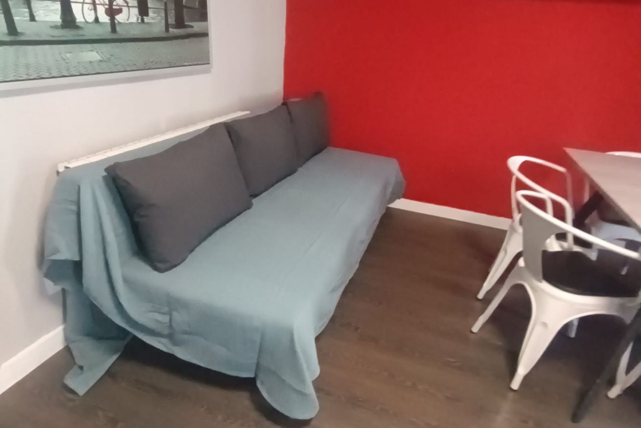 Una habitación con un sofá gris, sillas blancas, una pared roja y una ventana con vistas a árboles.