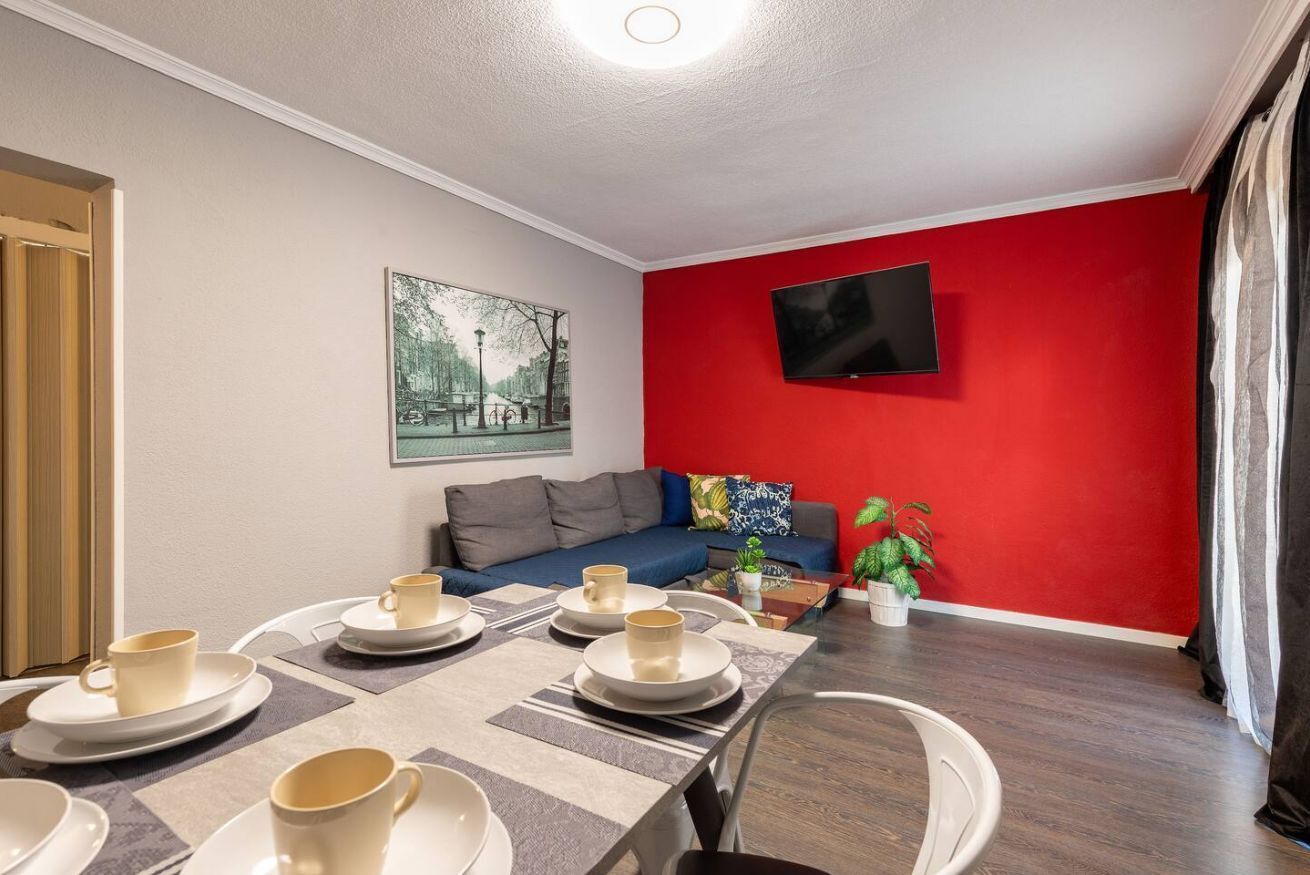 Ein modernes Wohnzimmer mit roter Akzentwand, einem Flachbildfernseher, einer Couch und einem Essbereich mit Tellern und Tassen.