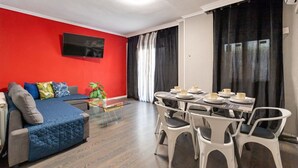 Un salon moderne avec un mur d’accent rouge, un canapé, un téléviseur, un coin repas avec des chaises et une table, et une plante.