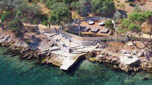 An aerial view of a coastal property with a stone bridge leading to a seating area with outdoor loungers, surrounded by trees and rocky terrain. - Exclusive Seafront Villa up to 18 – Breathtaking Views & Personal Chef & Daily maid Included! (Sparto)