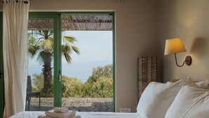 A hotel room with a bed, a lamp, a view of palm trees and the sea, and a wicker wall decoration. - Exclusive Seafront Villa up to 18 – Breathtaking Views & Personal Chef & Daily maid Included! (Sparto)