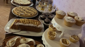 Een tafel met diverse desserts, waaronder taarten, gebakjes en cupcakes, met een witte tafelkleed en houten stoelen op de achtergrond.