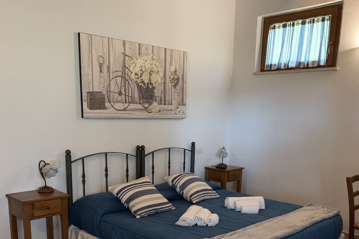 Een slaapkamer met een bed, nachtkastje, stoel, tafel en een raam met gordijnen.