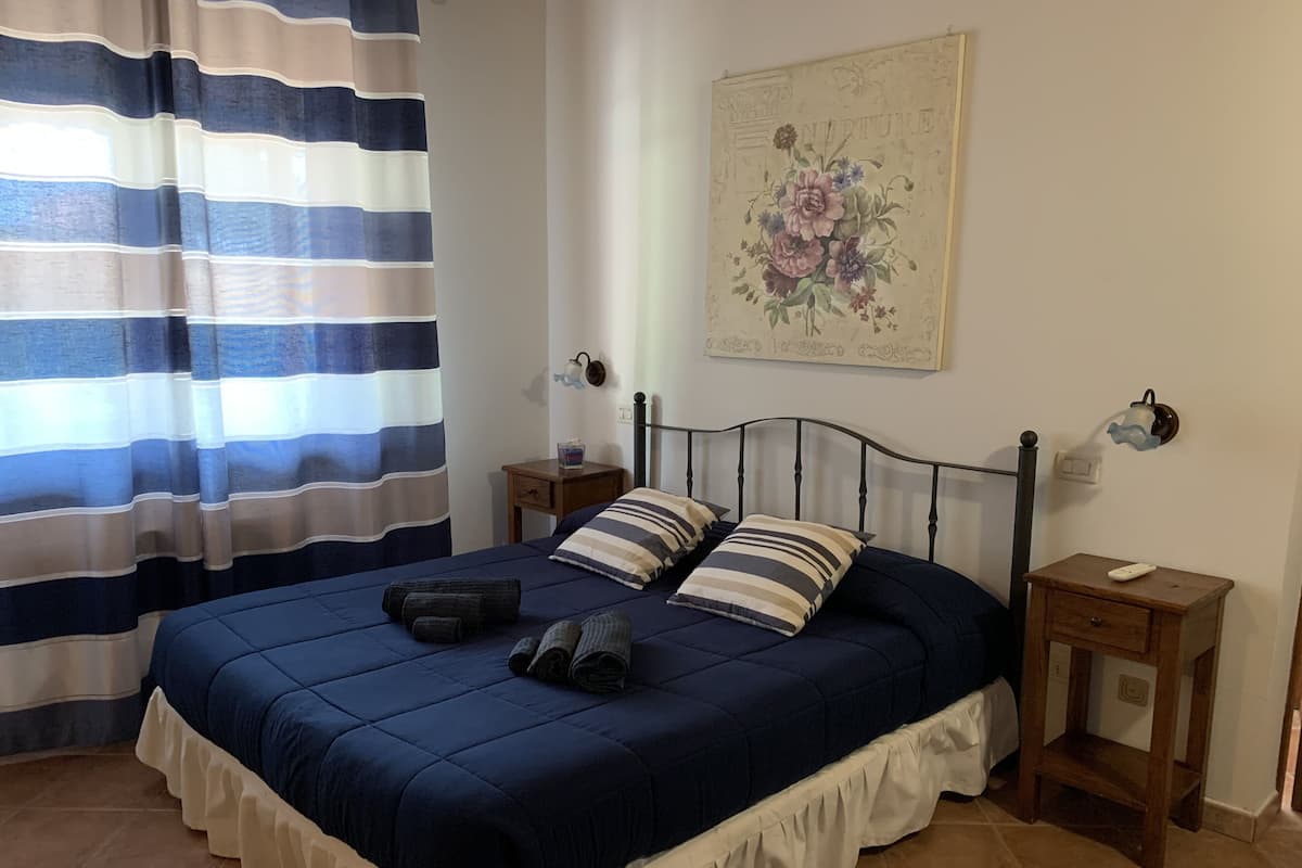 Een slaapkamer met een bed, gestreepte gordijnen, een nachtkastje, een lamp en een bloemig kunstwerk aan de muur.