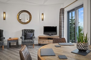 Un salon moderne avec une table ronde en bois, deux fauteuils, un téléviseur et une grande fenêtre donnant sur un paysage arboré.