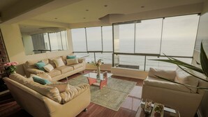 A modern living room with a large window overlooking the sea, furnished with beige sofas and a glass coffee table. - Exclusive oceanfront Penthouse in Miraflores with rooftop terrace (Urb Santa Cruz)