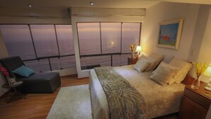 A bedroom with a large window offering a view of the sea, a bed with a patterned bedspread, a bedside table with a lamp, and a small side table with a vase. - Exclusive oceanfront Penthouse in Miraflores with rooftop terrace (Urb Santa Cruz)
