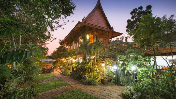 A traditional Thai house with a steeply pitched roof, surrounded by lush greenery and a paved pathway. - Cattani's Riverside Home (Ayutthaya)