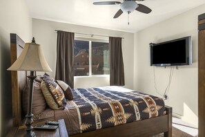 Une chambre à coucher avec un lit, un téléviseur fixé au mur, un ventilateur de plafond et une fenêtre avec des rideaux.