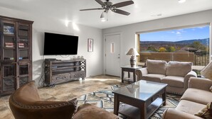 A living room with a flat-screen TV, a ceiling fan, a large window with a view of the mountains, and a cozy seating area. - Close to Town & National Parks | Pool & Hot Tub (Moab)