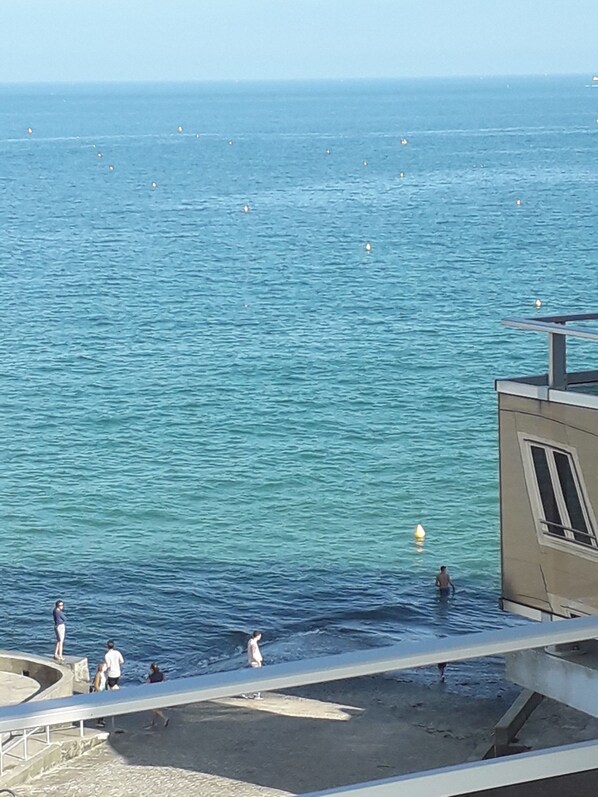 Vista costera con un edificio junto al mar, un muelle y gente cerca del agua.
