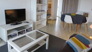 Uma sala de estar moderna com televisão de tela plana, prateleiras brancas, área de refeições com mesa e cadeiras, e um espaço de estar aconchegante com um sofá.
