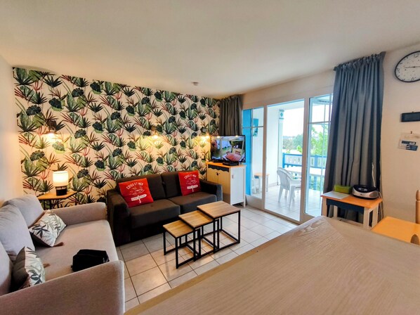 Hotellihuone, jossa on sohva, sänky, televisio, pieni pöytä ja parveke, jolta on näkymä.