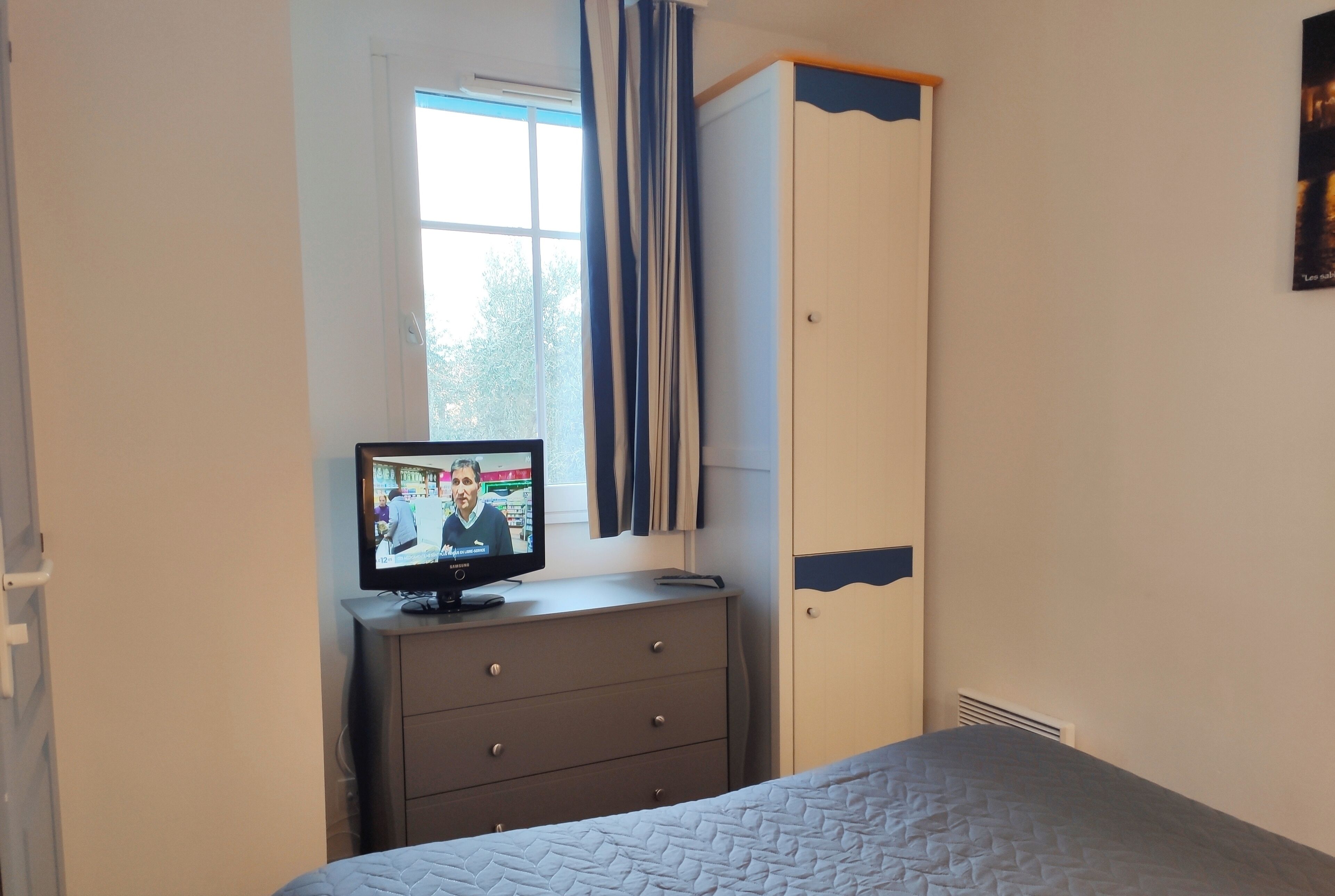 Un dormitorio con cama, cómoda, televisor encima de la cómoda, ventana con cortinas y armario.